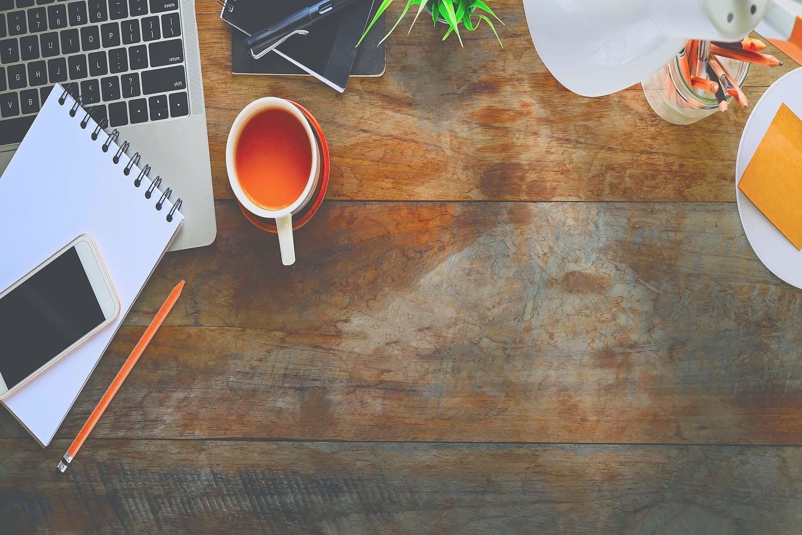 A wooden desk seen from above: a cup of tea, a phone lying on an open notebook, a pencil, and the corner of a laptop.