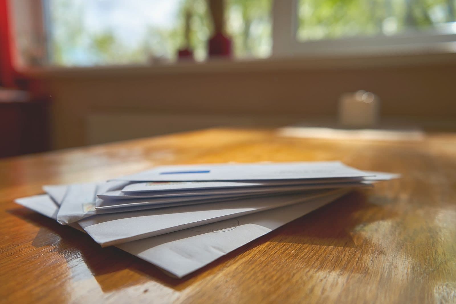 A stack of unopened envelopes on a wooden table by a window.