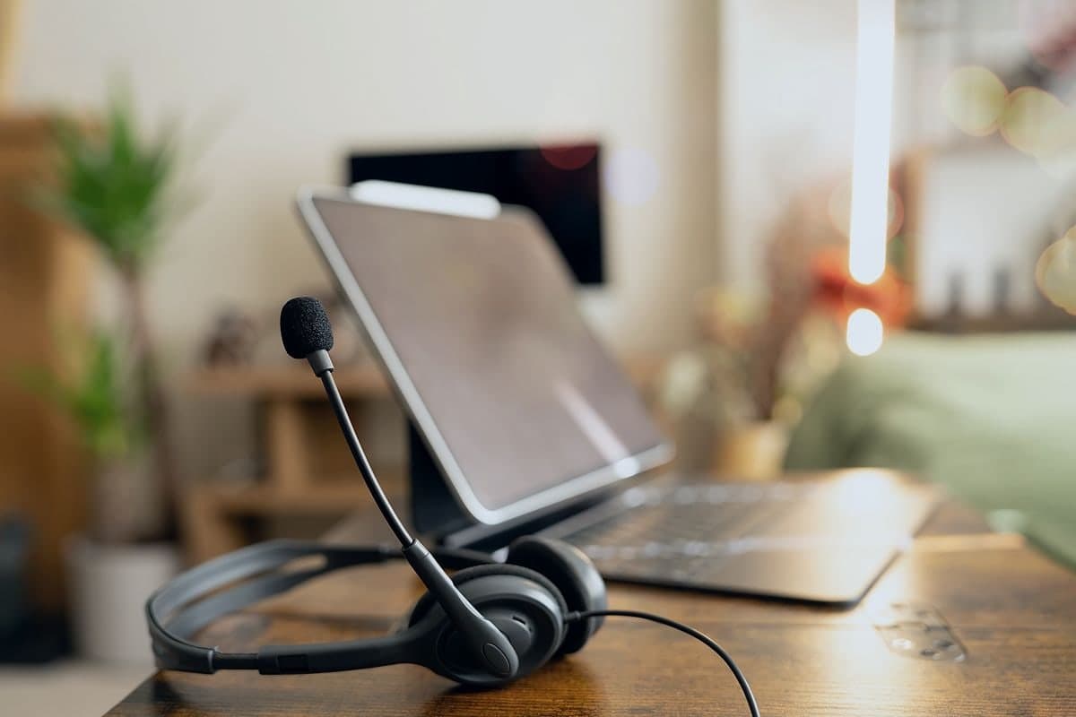 A headset resting on a wooden desk beside an open laptop, a lamp lit behind it.