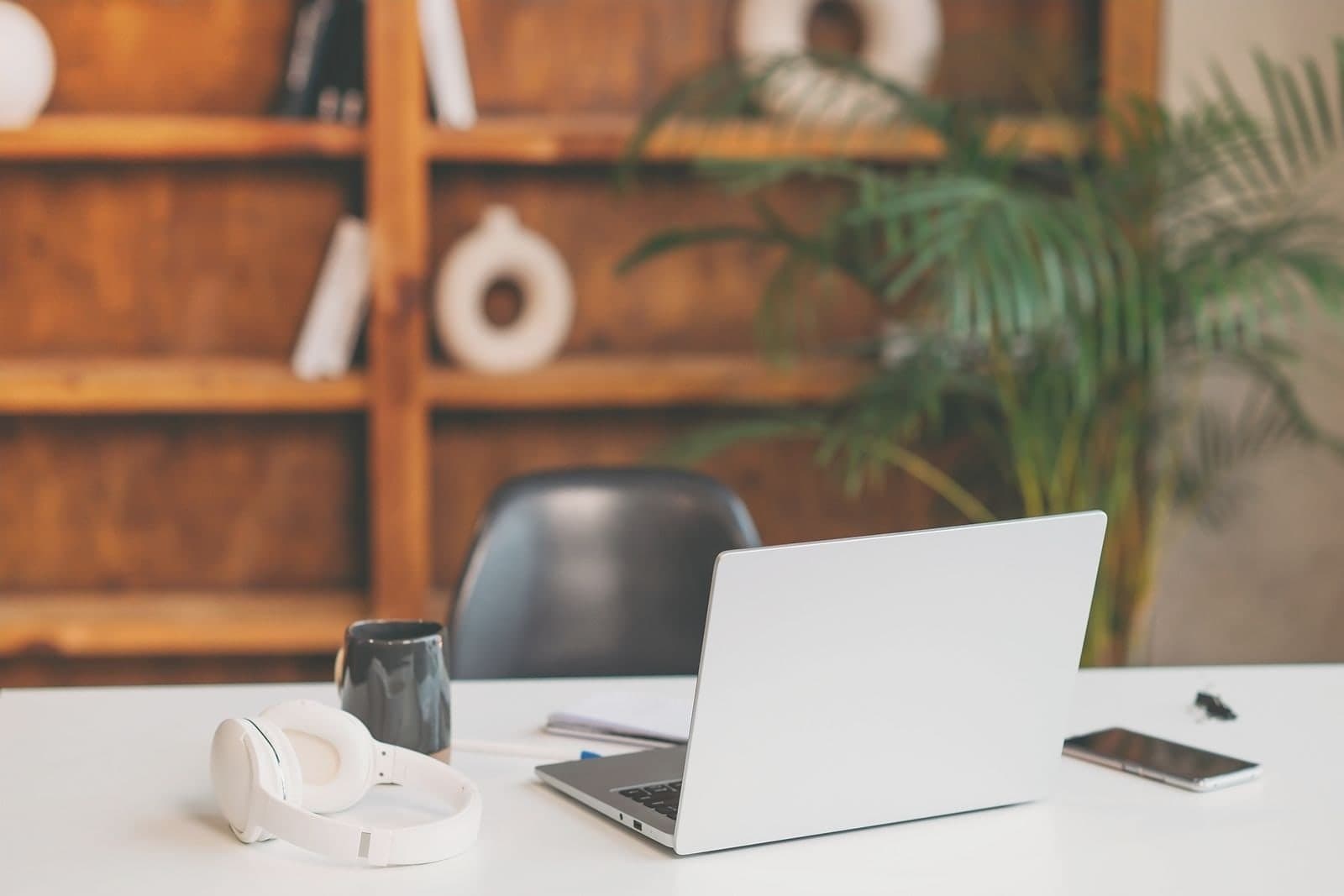 A white office desk with a laptop, a mug, headphones and a phone, shelves and a palm behind.