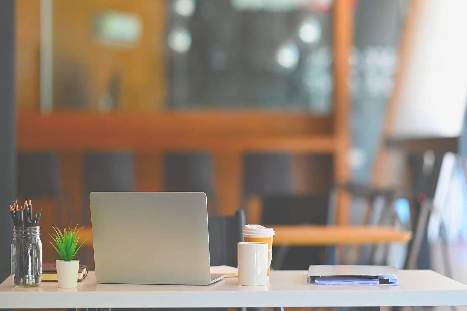 A laptop open on a desk by a window, a coffee cup beside it and the chair pushed back.