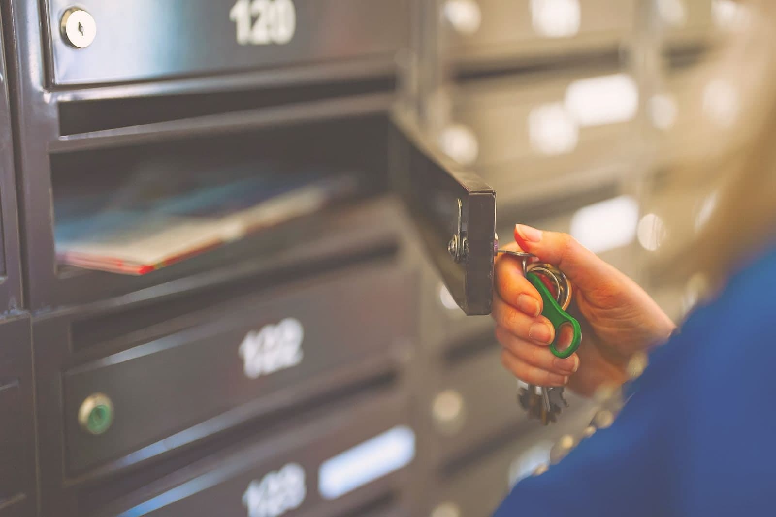 A hand unlocking one door in a bank of apartment mailboxes, post visible inside.