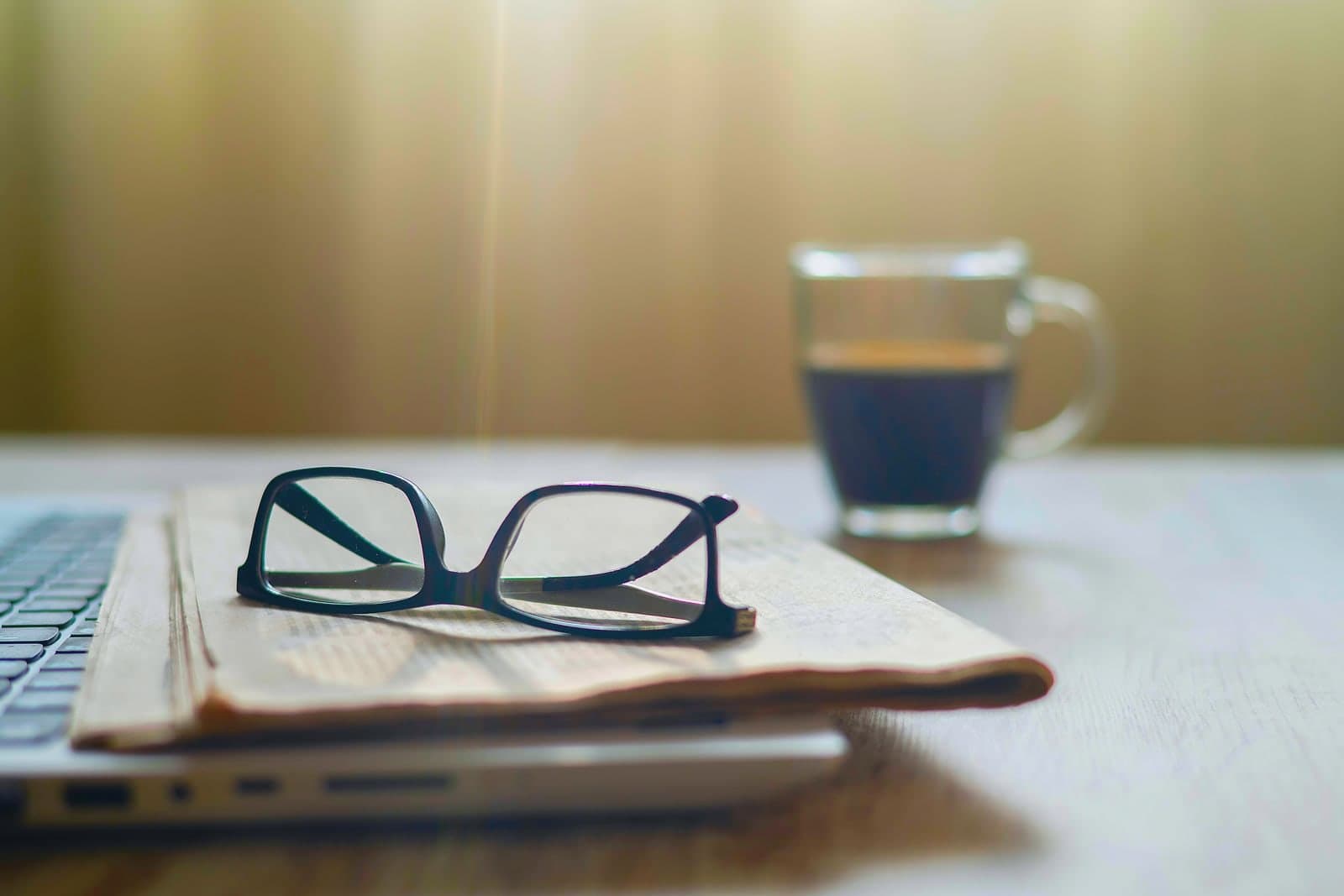 A folded newspaper and reading glasses on a closed laptop, morning coffee alongside.